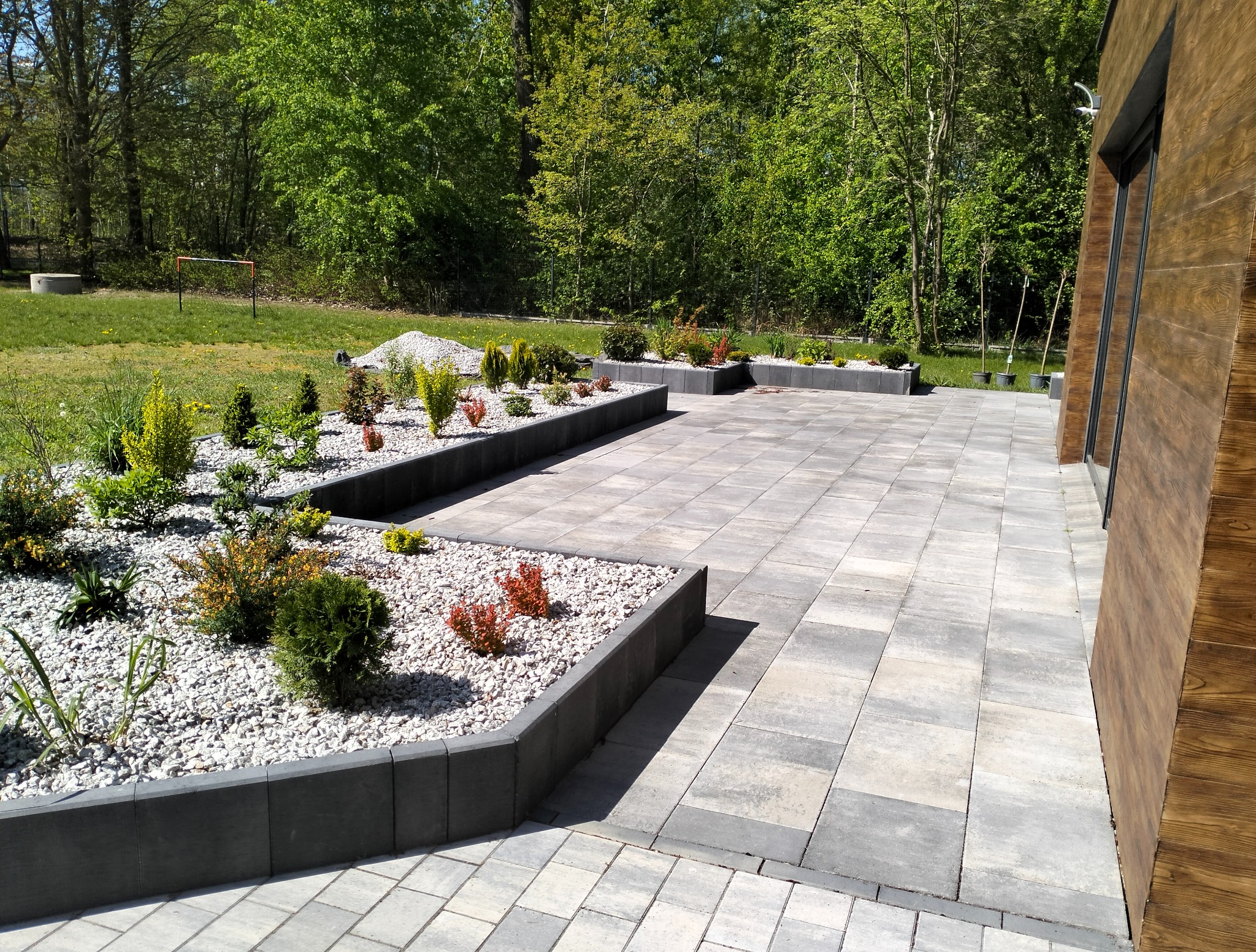 Szara kostka brukowa na podwórku z rabatami, obok budynku z drewnianą elewacją. W tle zieleń drzew i trawnik z bramką piłkarską.