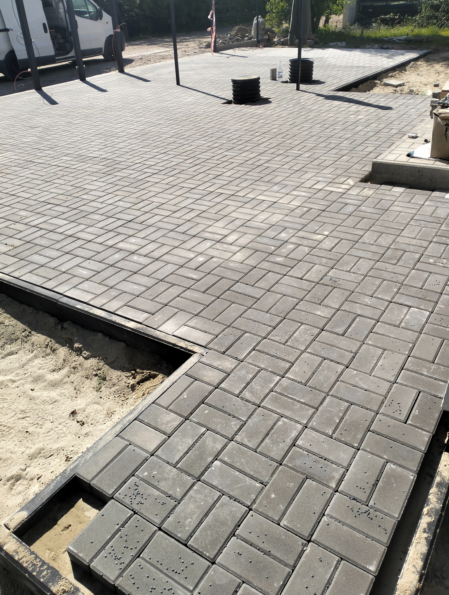 Układanie szarej kostki brukowej w jodełkę. Widoczne czarne obrzeża i piasek pod spodem. Częściowo ułożona nawierzchnia placu z elementami infrastruktury.