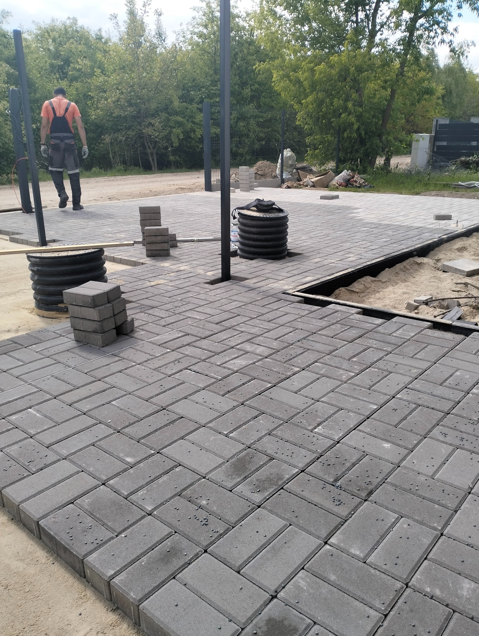 Szara kostka brukowa ułożona w jodełkę, widoczne studzienki kanalizacyjne i przygotowane podłoże pod dalszą część nawierzchni. Prace w toku, widoczna osoba w oddali.