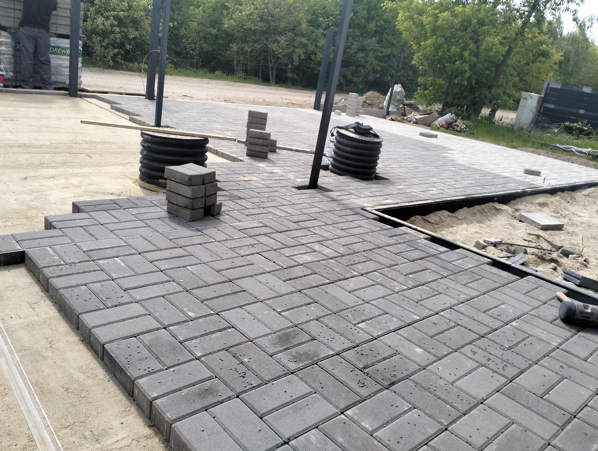Szara kostka brukowa ułożona we wzór jodełki, widoczne czarne słupki konstrukcji i przygotowane materiały. Częściowo ułożona nawierzchnia, piasek i obrzeża.