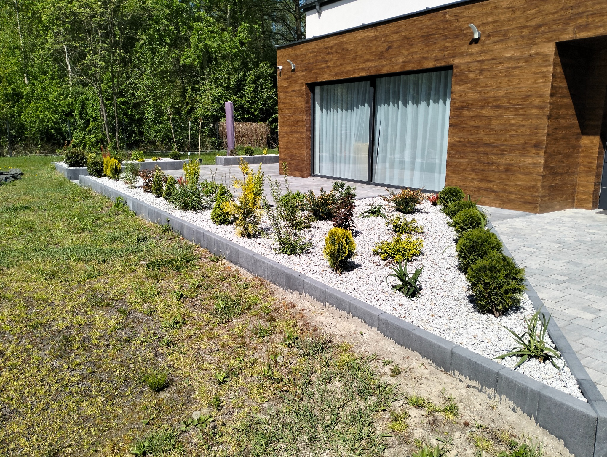 Nowoczesny ogród z obrzeżami z kostki, białym żwirem i różnorodną roślinnością na tle budynku z drewnianą elewacją i szarym brukiem.
