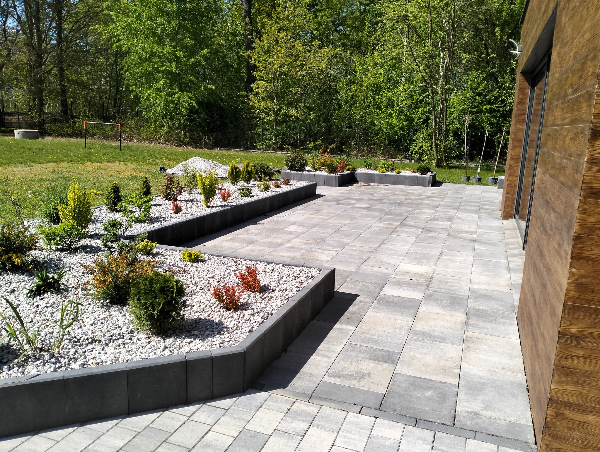 Ułożona kostka brukowa tworząca patio z rabatami obsadzonymi roślinami. Szara kostka, obrzeża z ciemnego betonu, jasne kamienie i zieleń na tle drzew.