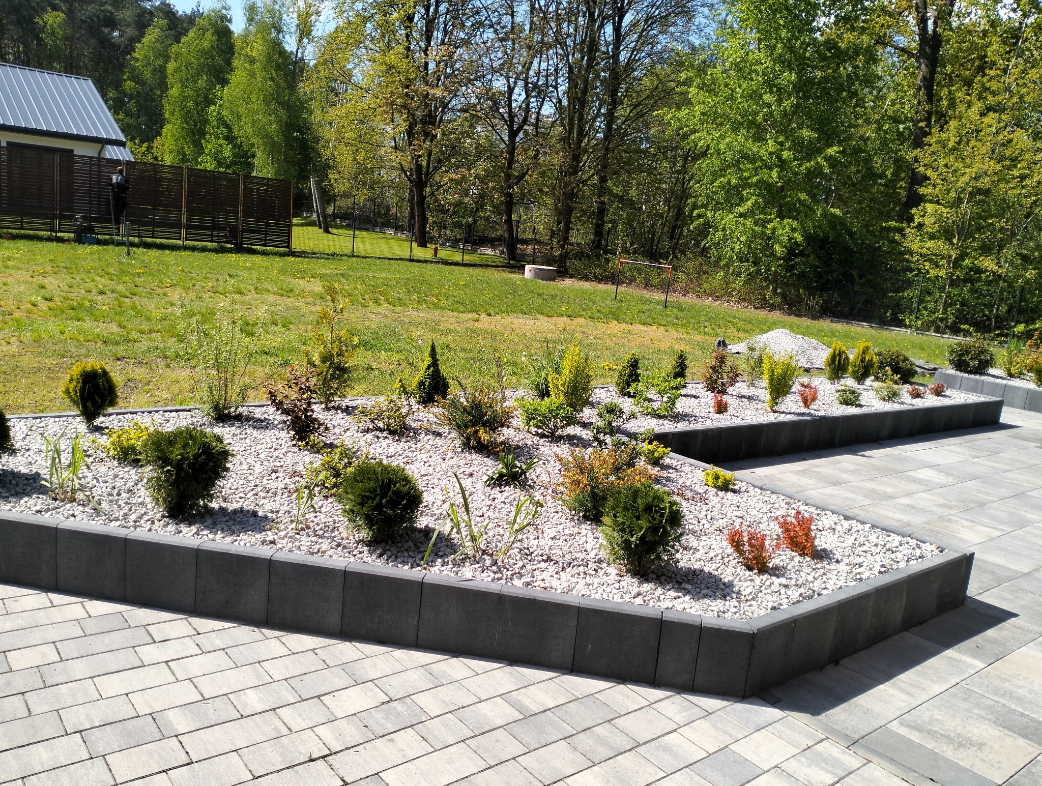 Ogród z rabatami wykończonymi szarą kostką, wypełnionymi białym grysem i różnorodną roślinnością. W tle trawnik i ogrodzenie z drewnianych paneli.