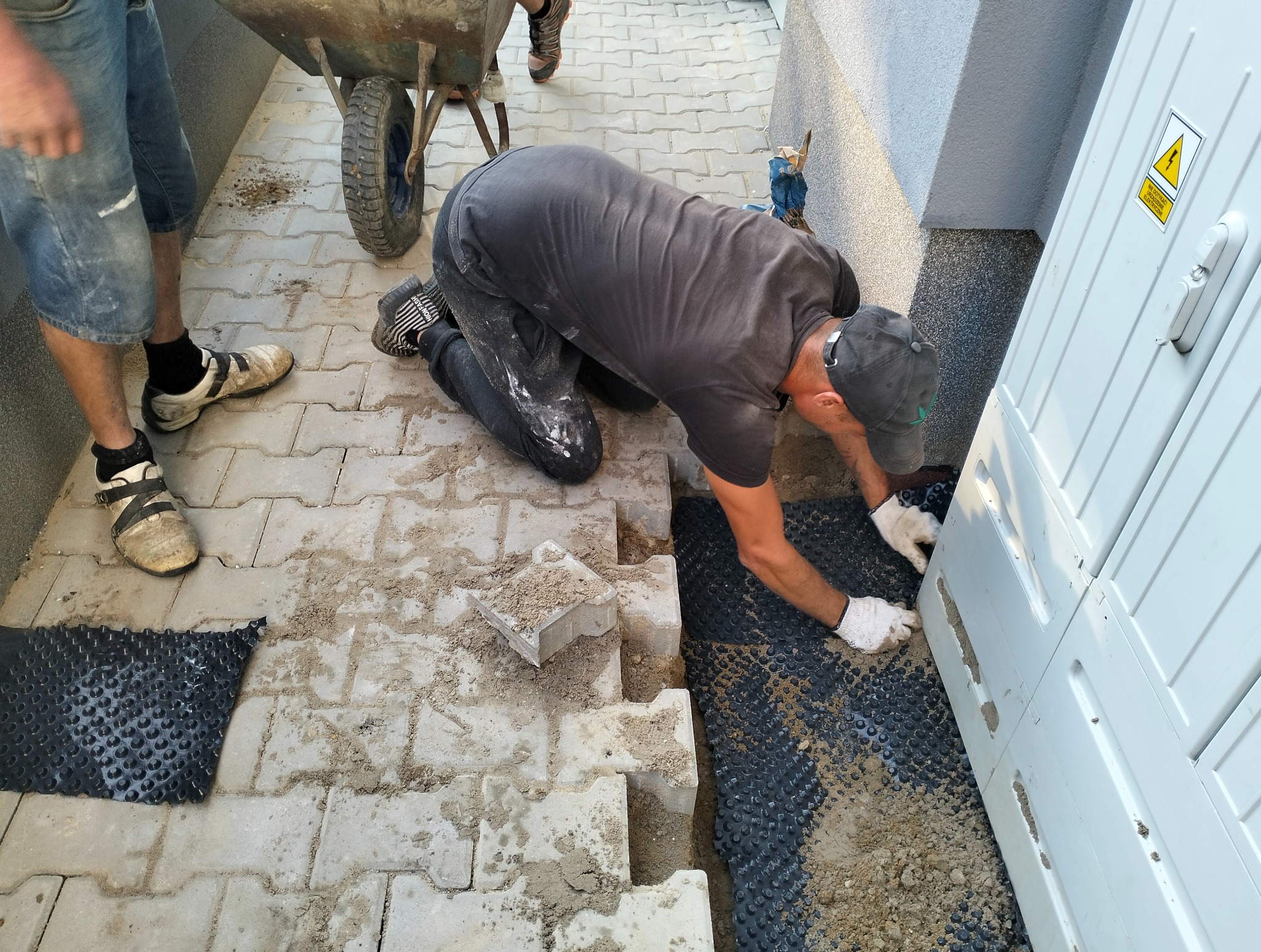 Mężczyzna w rękawicach układa kostkę brukową przy białej skrzynce elektrycznej, obok taczka i fragment ułożonej nawierzchni z kostki. Widoczne czarne podłoże.