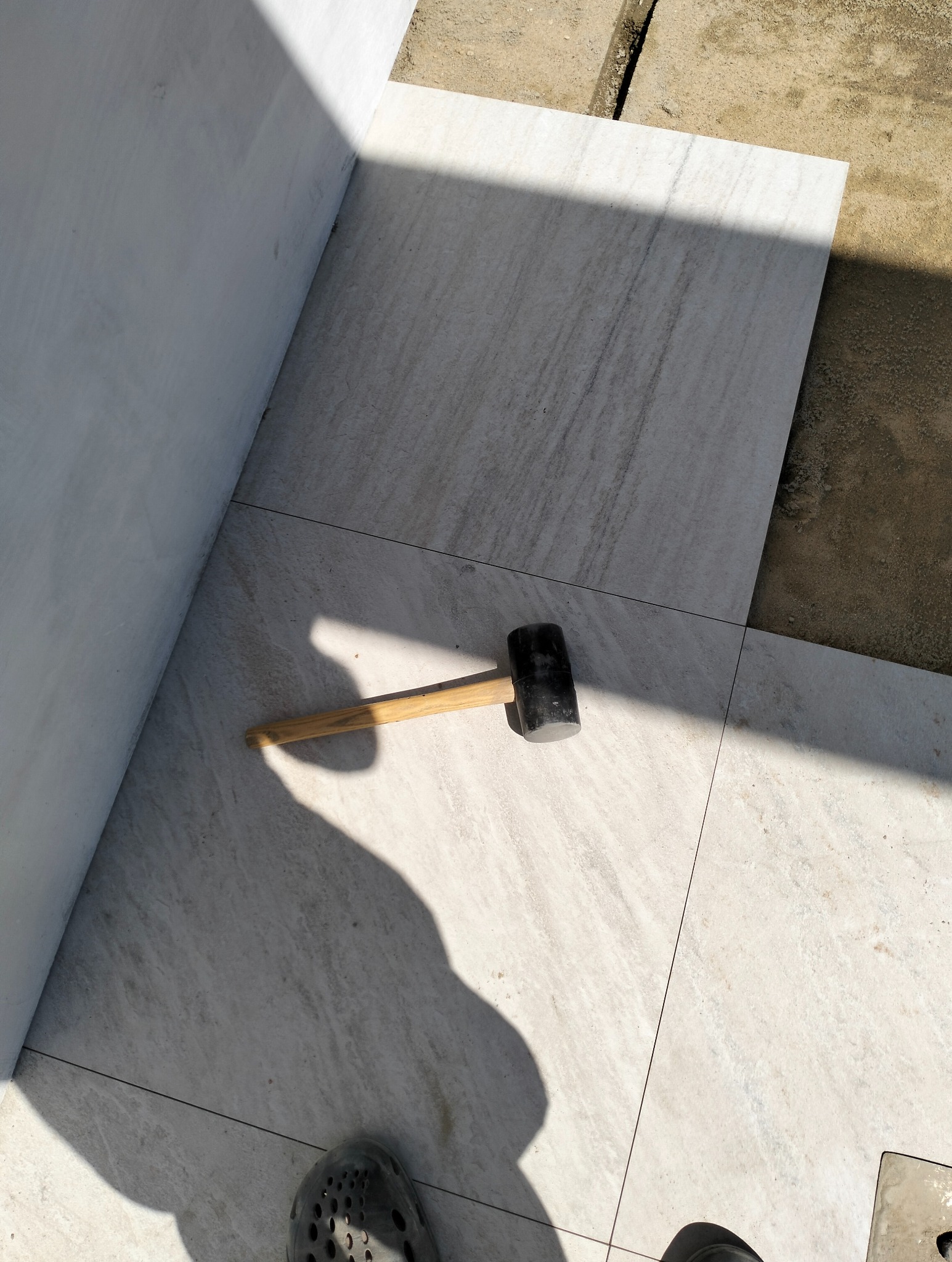 Układanie jasnych płyt chodnikowych przy ścianie. Gumowy młotek leży na powierzchni. Częściowo widoczne stopy w obuwiu roboczym.