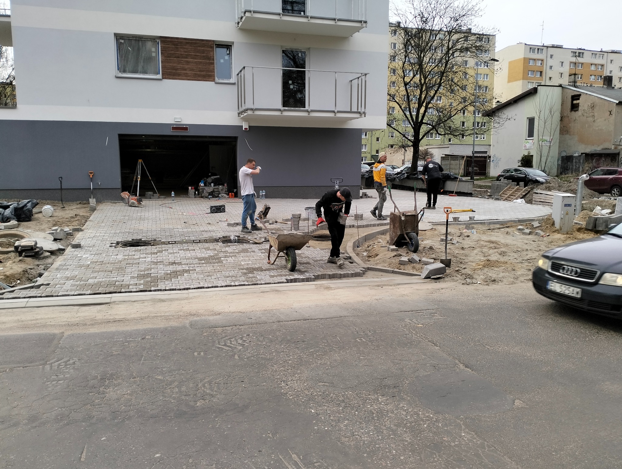 Praca wre: ekipa brukarzy układa szarą kostkę brukową na podjeździe przed nowoczesnym budynkiem z balkonami. W tle widać inne budynki mieszkalne i przejeżdżający samochód.
