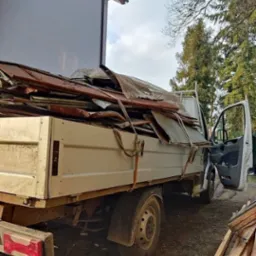 Ciężarówka wypełniona gruzem budowlanym i elementami dachu, zaparkowana na zewnątrz, z otwartymi drzwiami kierowcy, gotowa do wywozu odpadów.