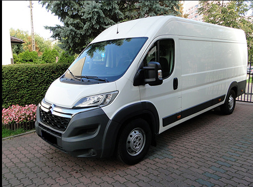 Biały furgon Citroen Jumper, zaparkowany na kostce brukowej, z widocznymi elementami zieleni w tle. Auto gotowe do transportu gruzu po usunięciu ścian.