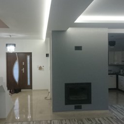 KM Rewers Remonty - Nowoczesne wnętrze domu po remoncie: szara ściana z kominkiem, oświetlenie LED sufitowe, widok na kuchnię i hol z brązowymi drzwiami. Parkiet w jasnych odcieniach.