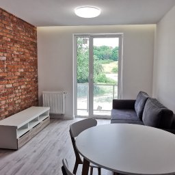 KM Rewers Remonty - Jasne wnętrze po remoncie: ceglana ściana, białe ściany, stół z krzesłami, sofa i szafka RTV. Widok na zieleń z balkonu.