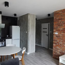 KM Rewers Remonty - Nowoczesne wnętrze: kuchnia z czarnym marmurem, ceglana ściana, okrągły stół i szare panele podłogowe. Widoczne oświetlenie sufitowe i biała lodówka.
