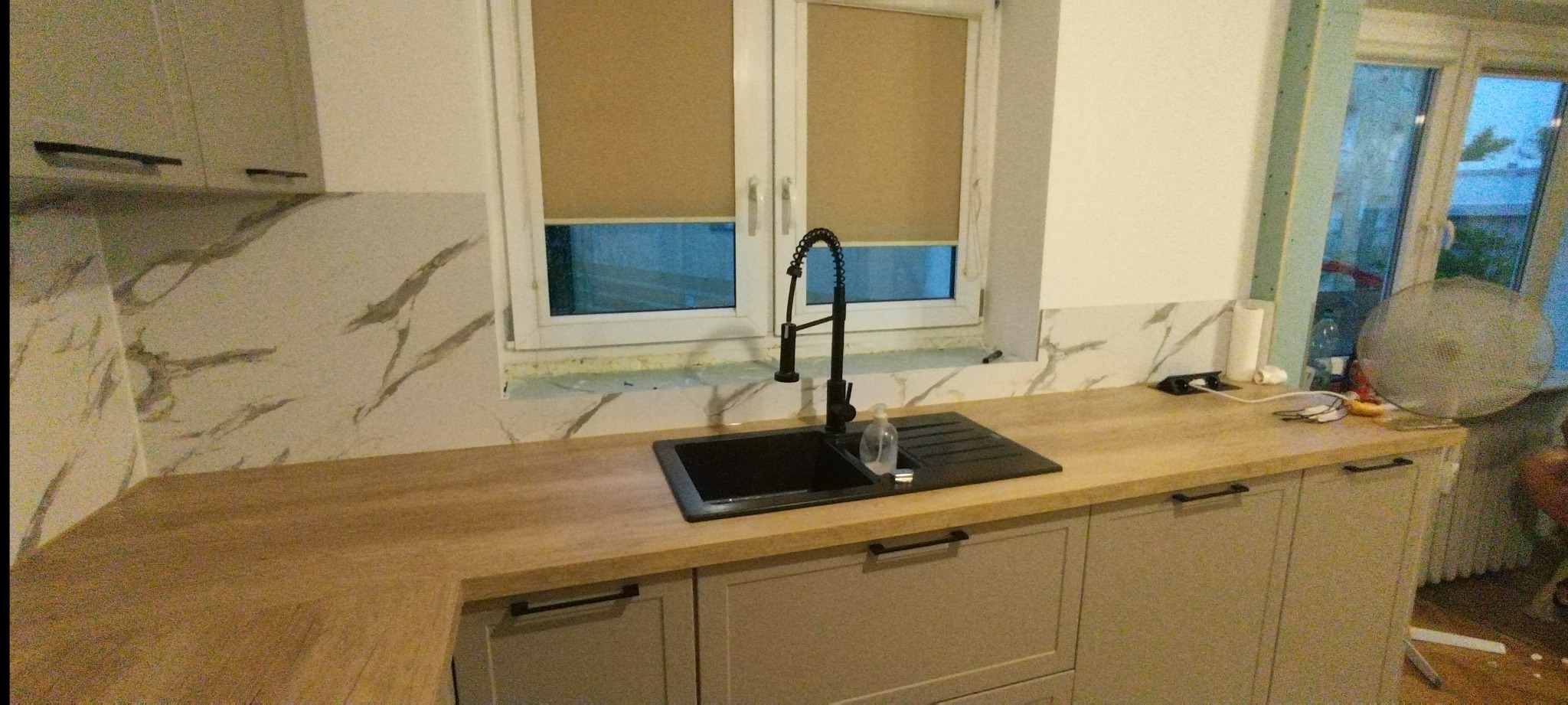 Nowoczesna kuchnia po remoncie: jasne szafki, drewniany blat, czarny zlew i bateria, marmurowe płytki na ścianie, roleta w oknie. Widok na fragment pomieszczenia.