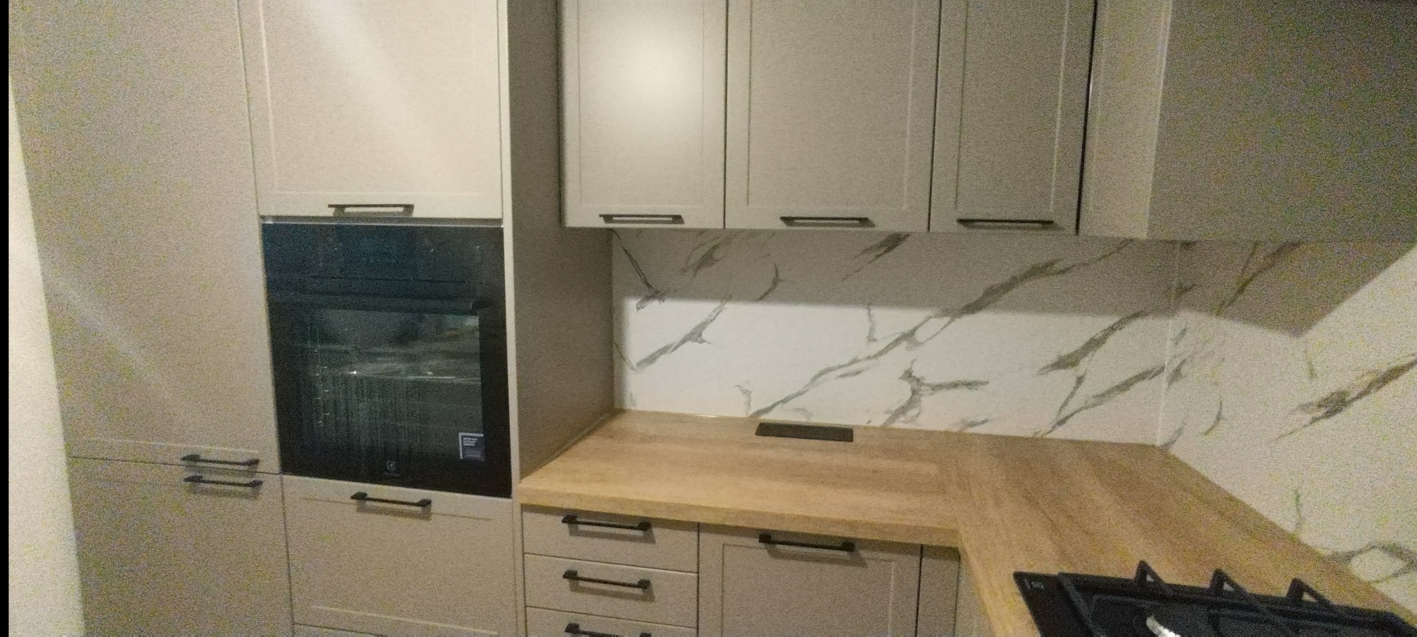 Jasna zabudowa kuchenna z piekarnikiem i marmurowym panelem nad blatem. Minimalistyczne uchwyty, ciepłe oświetlenie. Fragment płyty gazowej w prawym dolnym rogu.