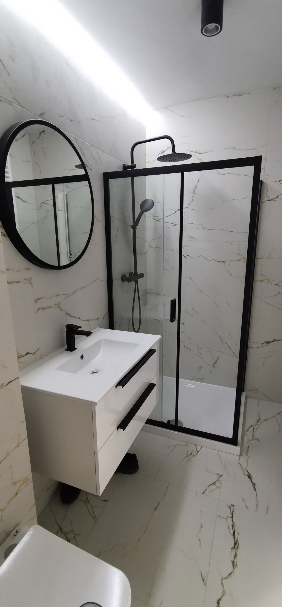 Nowoczesna łazienka z kabiną prysznicową typu walk-in, białą szafką z umywalką i okrągłym lustrem w czarnej ramie. Marmurowe płytki na ścianach i podłodze.