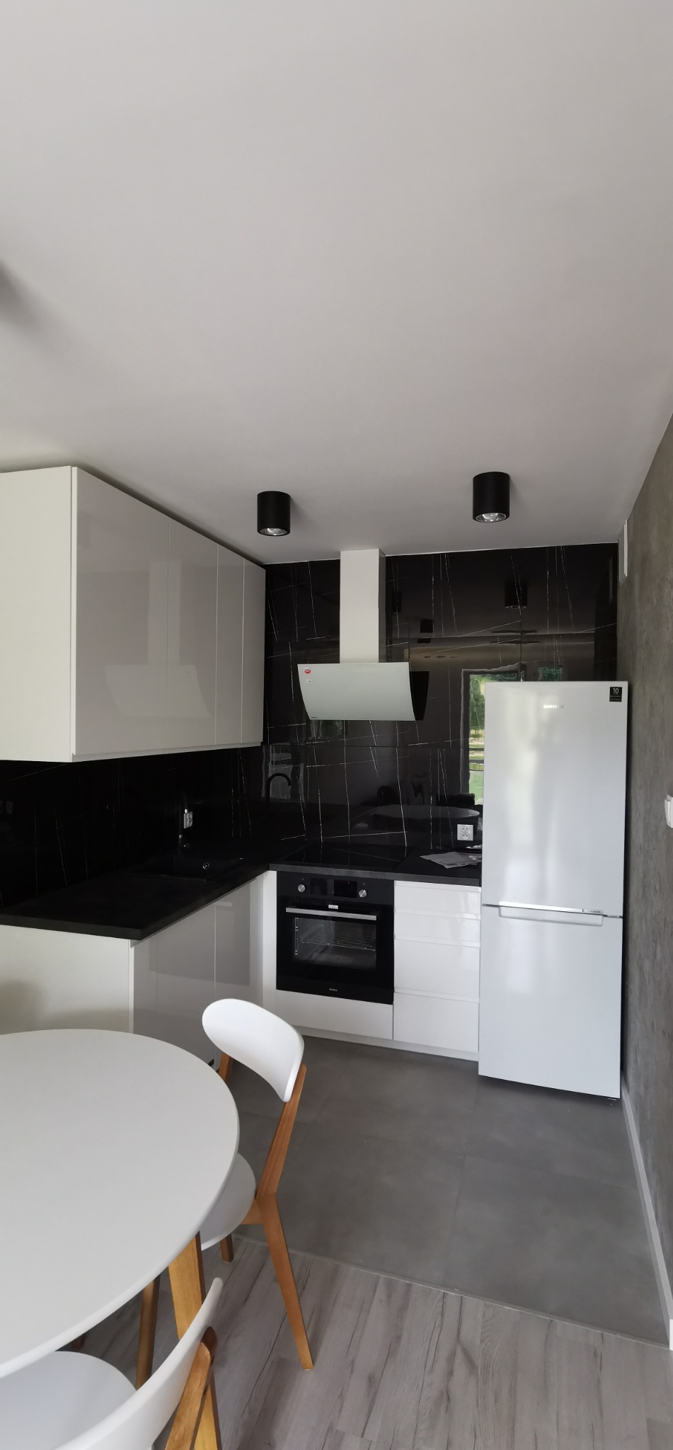 Nowoczesna kuchnia w bieli i czerni: białe szafki, czarny blat i backsplash z marmurowym wzorem. Okrągły stół z krzesłami w rogu. Minimalistyczne oświetlenie.