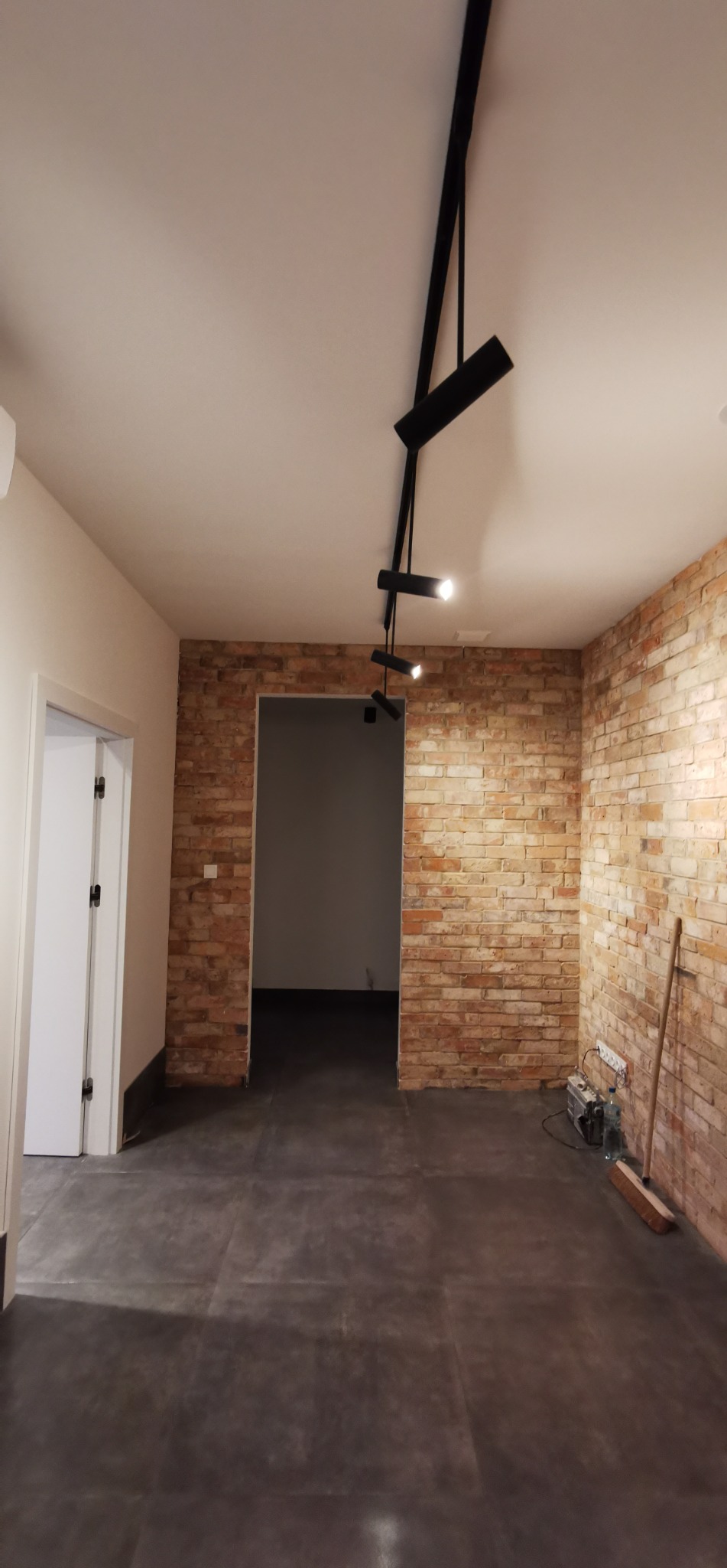 Surowe wykończenie wnętrza: ceglana ściana, ciemna podłoga, nowoczesne oświetlenie. Widoczne narzędzia i materiały budowlane, sugerujące etap prac wykończeniowych.