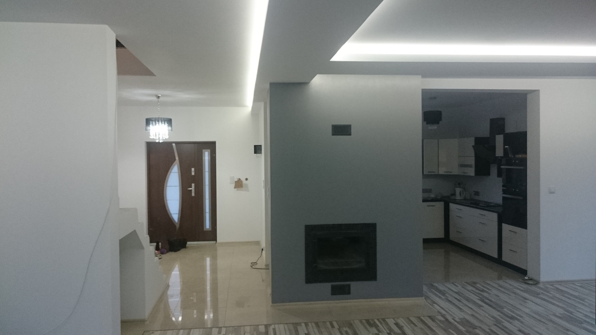 Nowoczesne wnętrze domu po remoncie: szara ściana z kominkiem, oświetlenie LED sufitowe, widok na kuchnię i hol z brązowymi drzwiami. Parkiet w jasnych odcieniach.