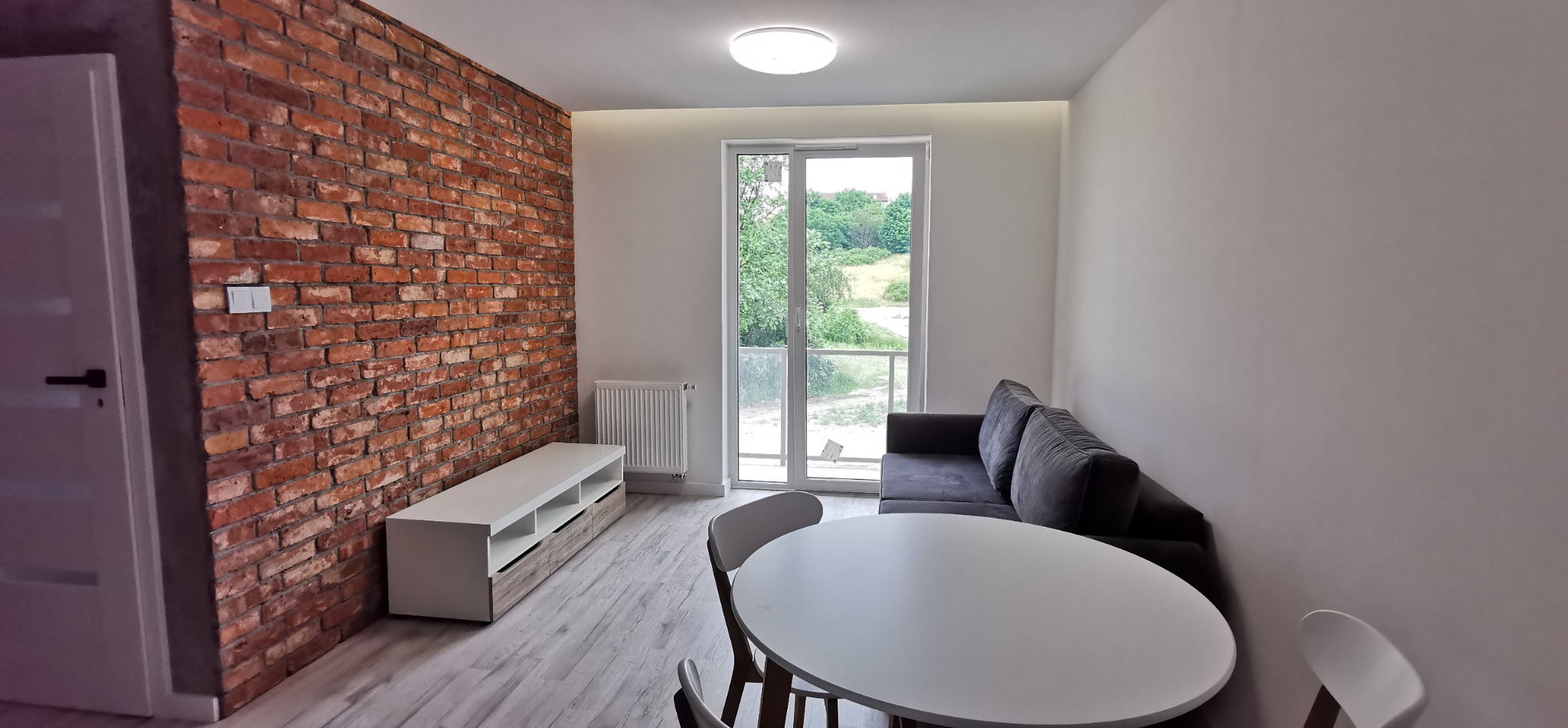 Jasne wnętrze po remoncie: ceglana ściana, białe ściany, stół z krzesłami, sofa i szafka RTV. Widok na zieleń z balkonu.