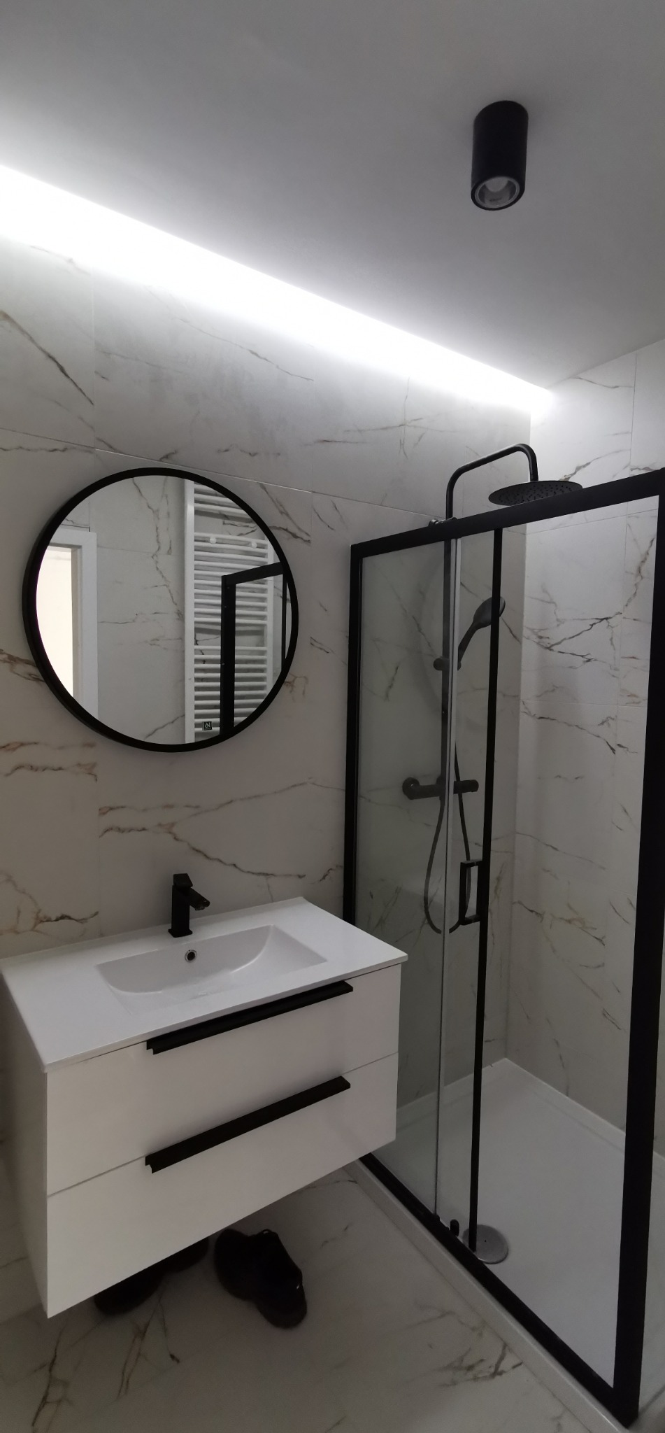 Nowoczesna łazienka z kabiną prysznicową typu walk-in, okrągłym lustrem i białą szafką pod umywalkę z czarnymi uchwytami na tle marmurowych płytek.