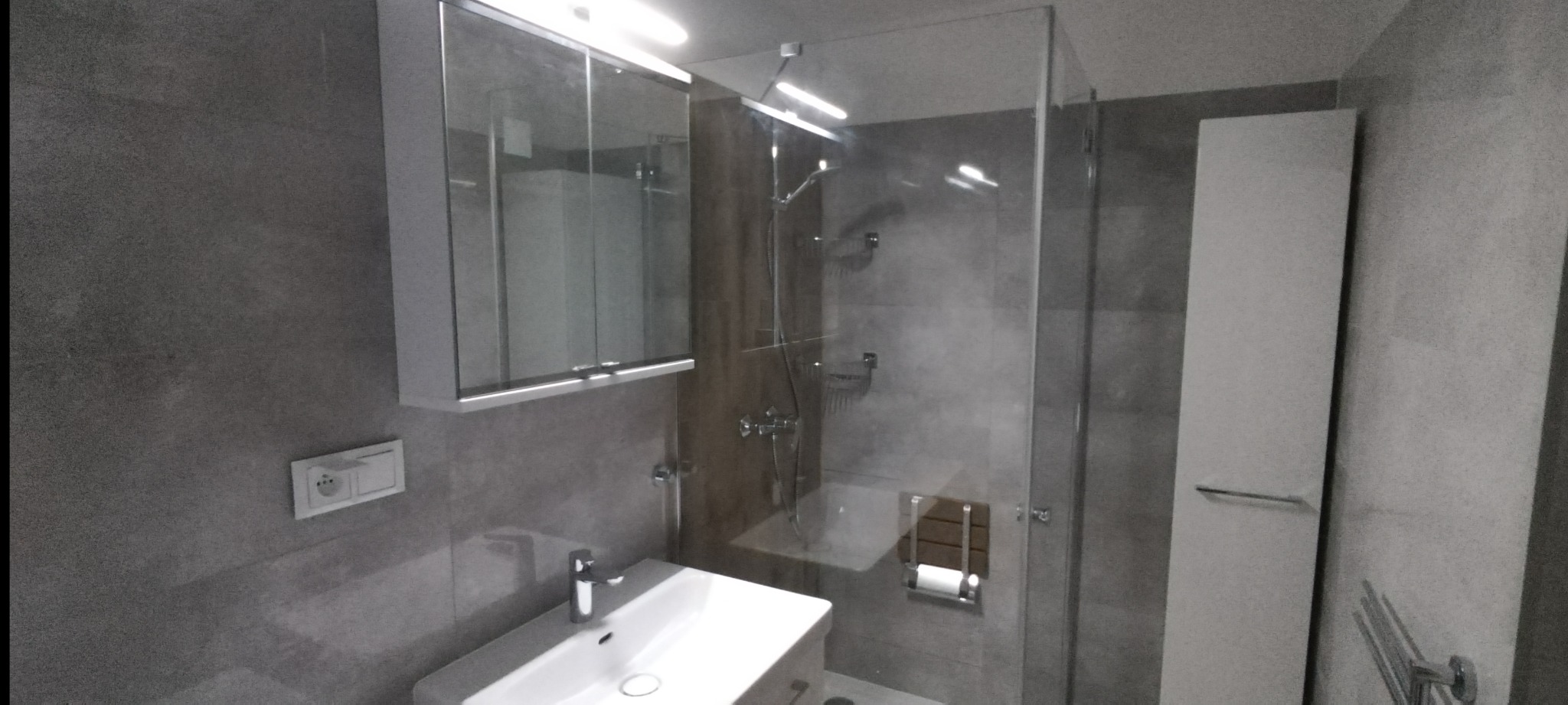 Nowoczesna łazienka z szarą glazurą, kabiną prysznicową ze szklanymi drzwiami, umywalką z szafką i lustrem z oświetleniem. Widoczny również biały słupek z relingiem.