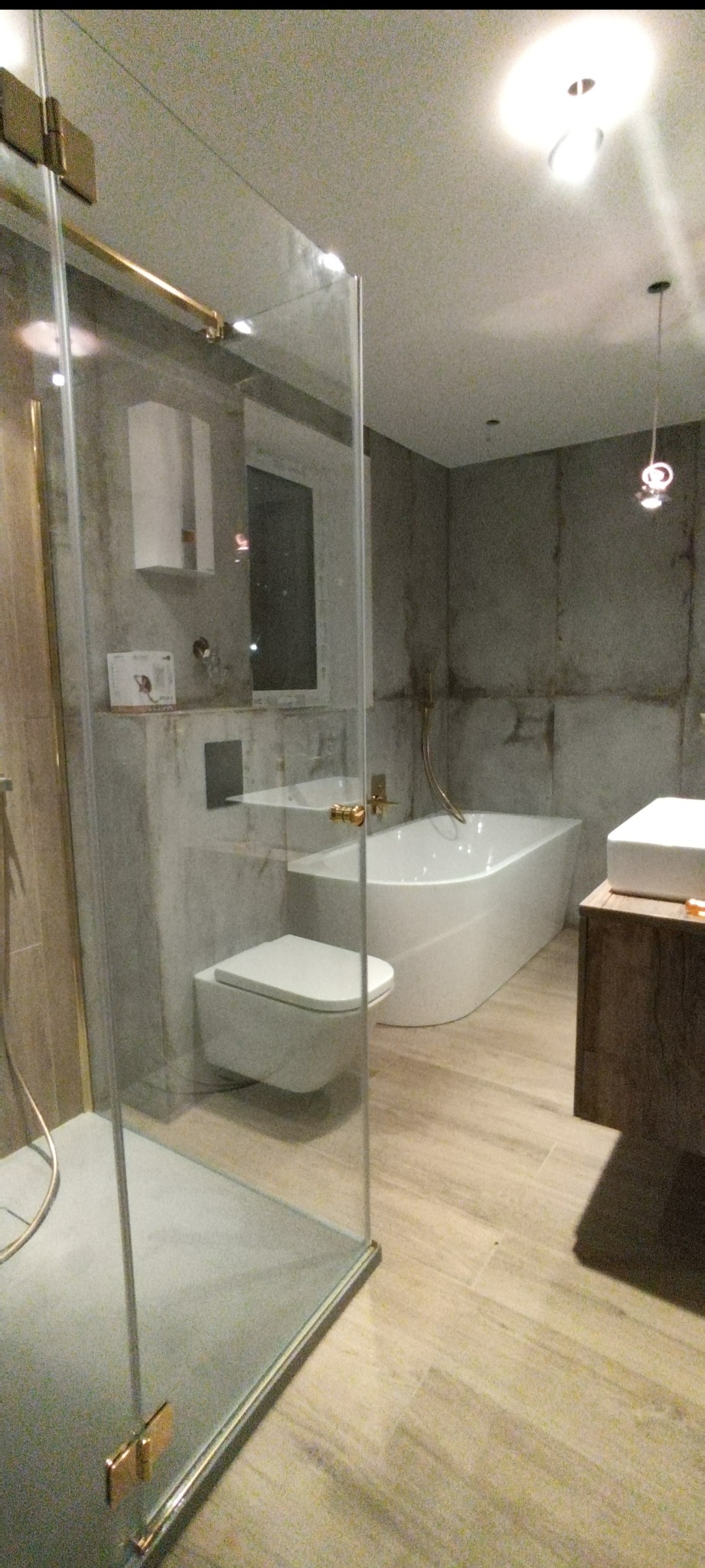 Nowoczesna łazienka z kabiną prysznicową ze złotymi detalami, wiszącą toaletą i wanną. Ściany wykończone w szarościach, drewnopodobna podłoga.