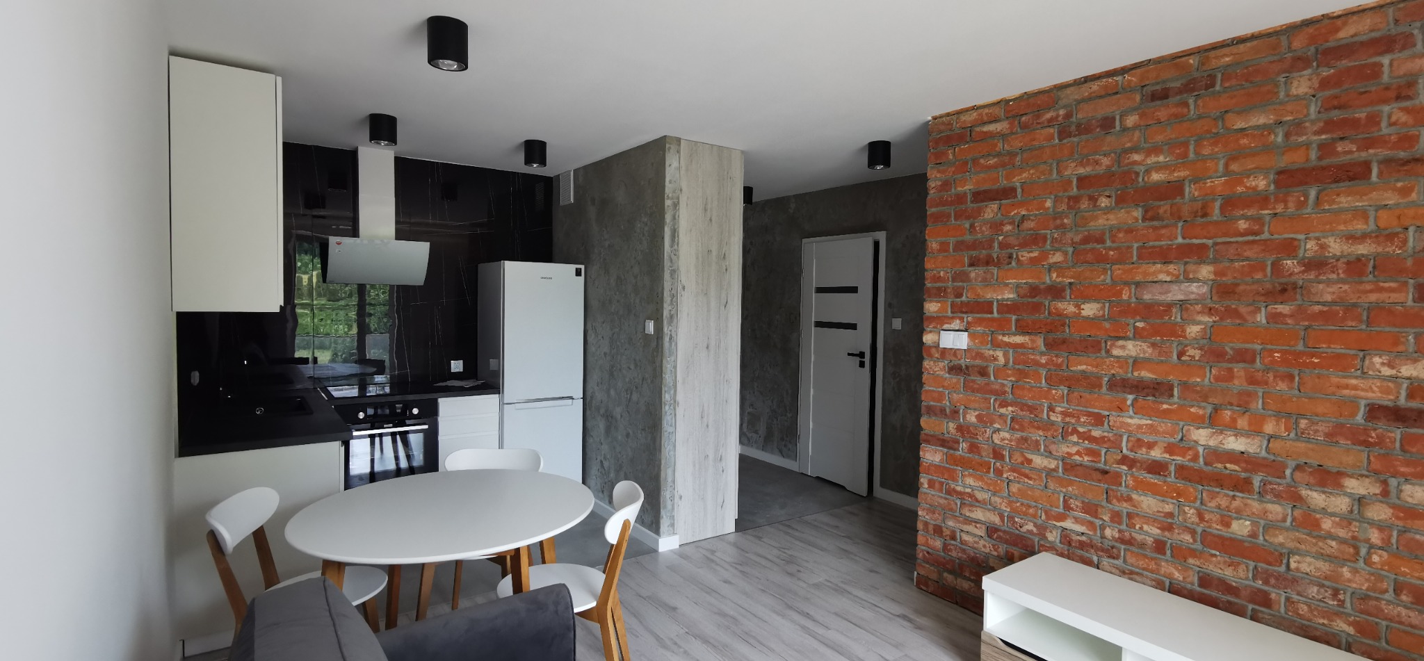 Nowoczesne wnętrze: kuchnia z czarnym marmurem, ceglana ściana, okrągły stół i szare panele podłogowe. Widoczne oświetlenie sufitowe i biała lodówka.