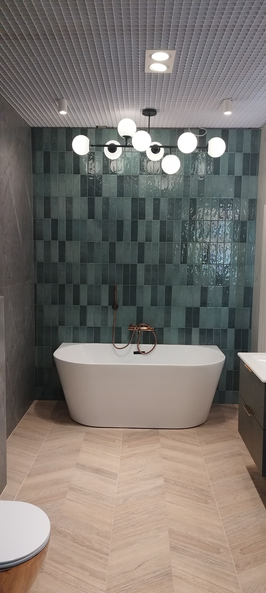Nowoczesna łazienka z wolnostojącą wanną na tle ściany z zielonej glazury i podłogi w jodełkę. Nad wanną designerska lampa.
