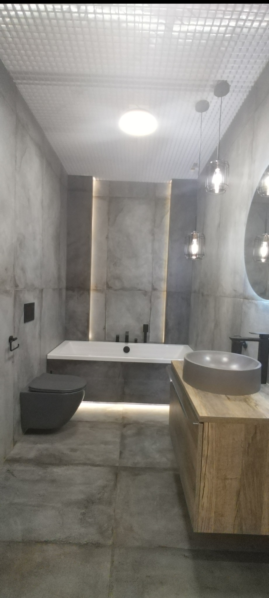 Nowoczesna łazienka w szarościach: podwieszana toaleta, wanna z podświetleniem LED, umywalka nablatowa na drewnianej szafce i designerskie lampy.