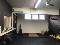 Klimatyzator ścienny w jasnym pomieszczeniu siłowni z czarnymi ścianami. Widoczne okna, sprzęt do ćwiczeń i podłoga z gumy.