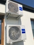 Dwa białe klimatyzatory Haier zamontowane na jasnej ścianie budynku, widoczne z boku, z fragmentem okna i dachu. Instalacja klimatyzacji zewnętrznej.