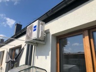 Biała jednostka zewnętrzna klimatyzacji Haier zamontowana na elewacji budynku, widoczna z dołu pod kątem, z oknami w tle i fragmentem dachu.