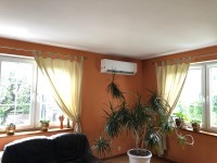 Biały klimatyzator ścienny w pomarańczowym pokoju z dwoma oknami i rośliną doniczkową. Widok z dołu, w tle fragment sofy.