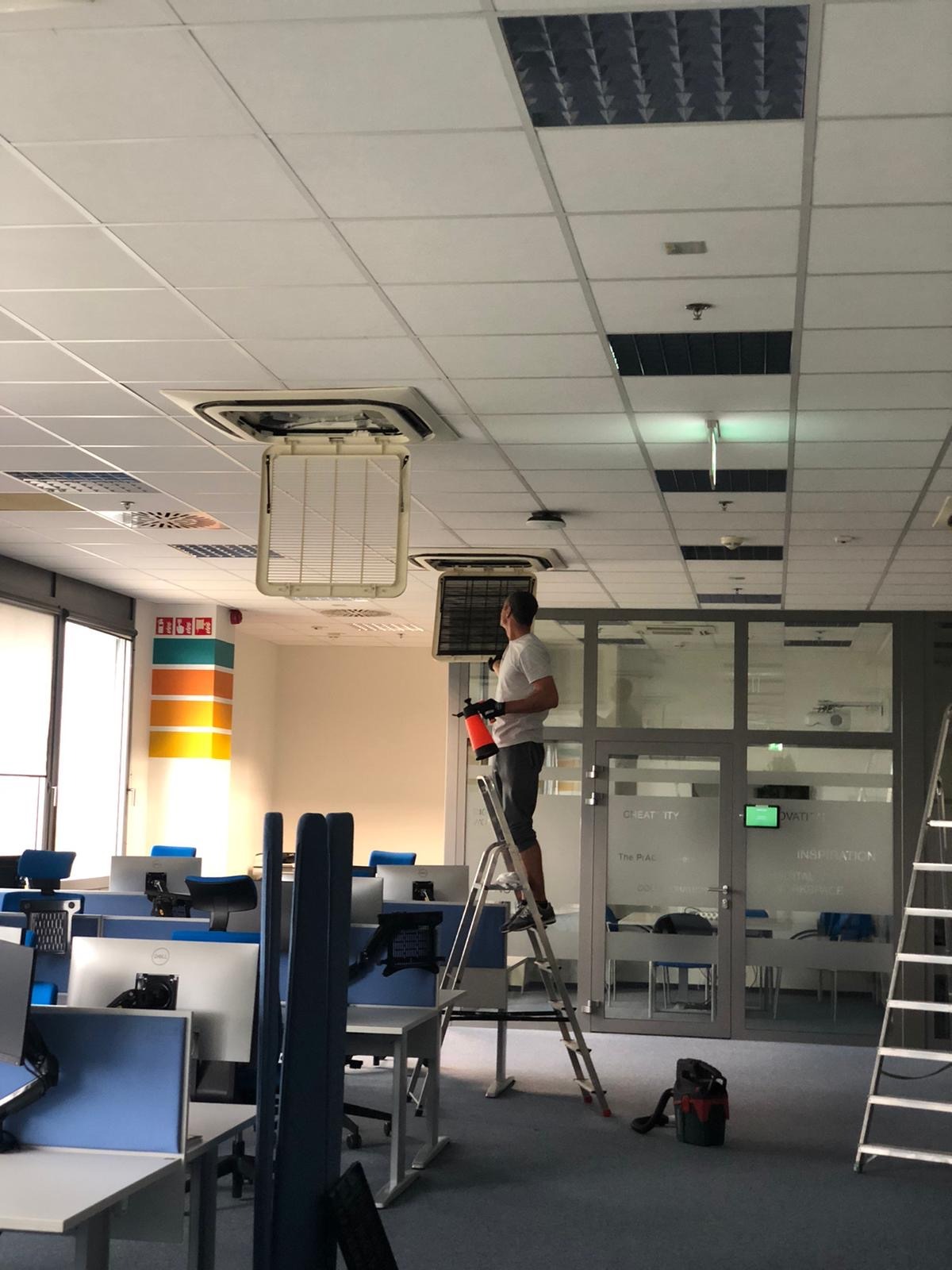Serwisant na drabinie czyści klimatyzację kasetonową w biurze. Widoczne biurka z komputerami, okna i inne klimatyzatory w suficie podwieszanym.