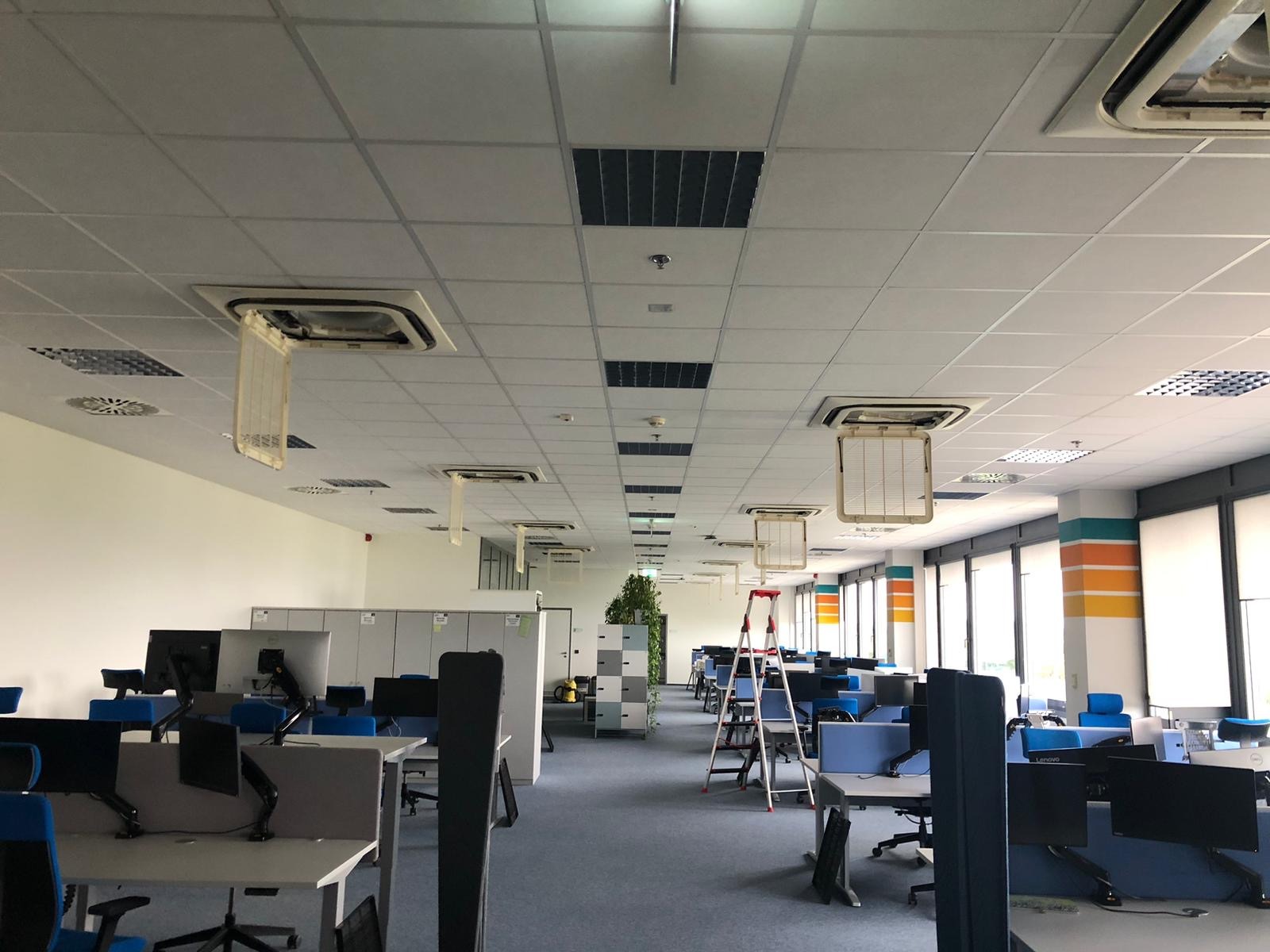Biuro po serwisie klimatyzacji: otwarte panele klimatyzatorów w suficie podwieszanym, drabina, biurka z komputerami. Widok na przestrzeń biurową.