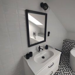 Remonty wykończenia wnętrz - Biała łazienka na poddaszu z umywalką na szafce, czarną armaturą i lustrem w czarnej ramie. W odbiciu widać wannę i okno dachowe. Uwagę przykuwają płytki podłogowe.