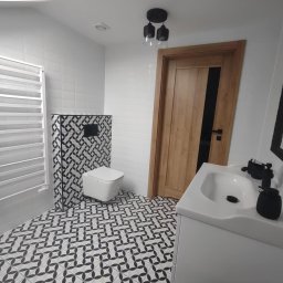 Remonty wykończenia wnętrz - Nowoczesna łazienka z białą umywalką, toaletą i grzejnikiem. Wzór geometryczny na podłodze i obudowie toalety. Drewniane drzwi z czarnym akcentem.