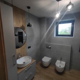 Remonty wykończenia wnętrz - Nowoczesna łazienka w szarościach z drewnianymi akcentami. Umywalka nablatowa, okrągłe lustro, podwieszana toaleta i bidet. Oświetlenie punktowe i drewnopodobna podłoga.