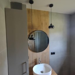 Remonty wykończenia wnętrz - Stylowa łazienka po remoncie: szara szafka z umywalką, okrągłe lustro i drewnopodobne panele ścienne, oświetlone dwoma czarnymi lampami wiszącymi.