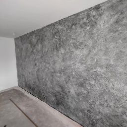 Remonty wykończenia wnętrz - Ściana z efektem dekoracyjnym w odcieniach szarości, surowa faktura, widoczne nierówności i cieniowania. Podłoga surowa, przygotowana do dalszych prac wykończeniowych.