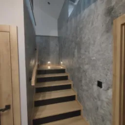 Nowoczesne schody z drewnianymi stopniami i szarą ścianą o fakturze betonu, oświetlone punktowymi lampami. Widok z dołu na górę klatki schodowej.