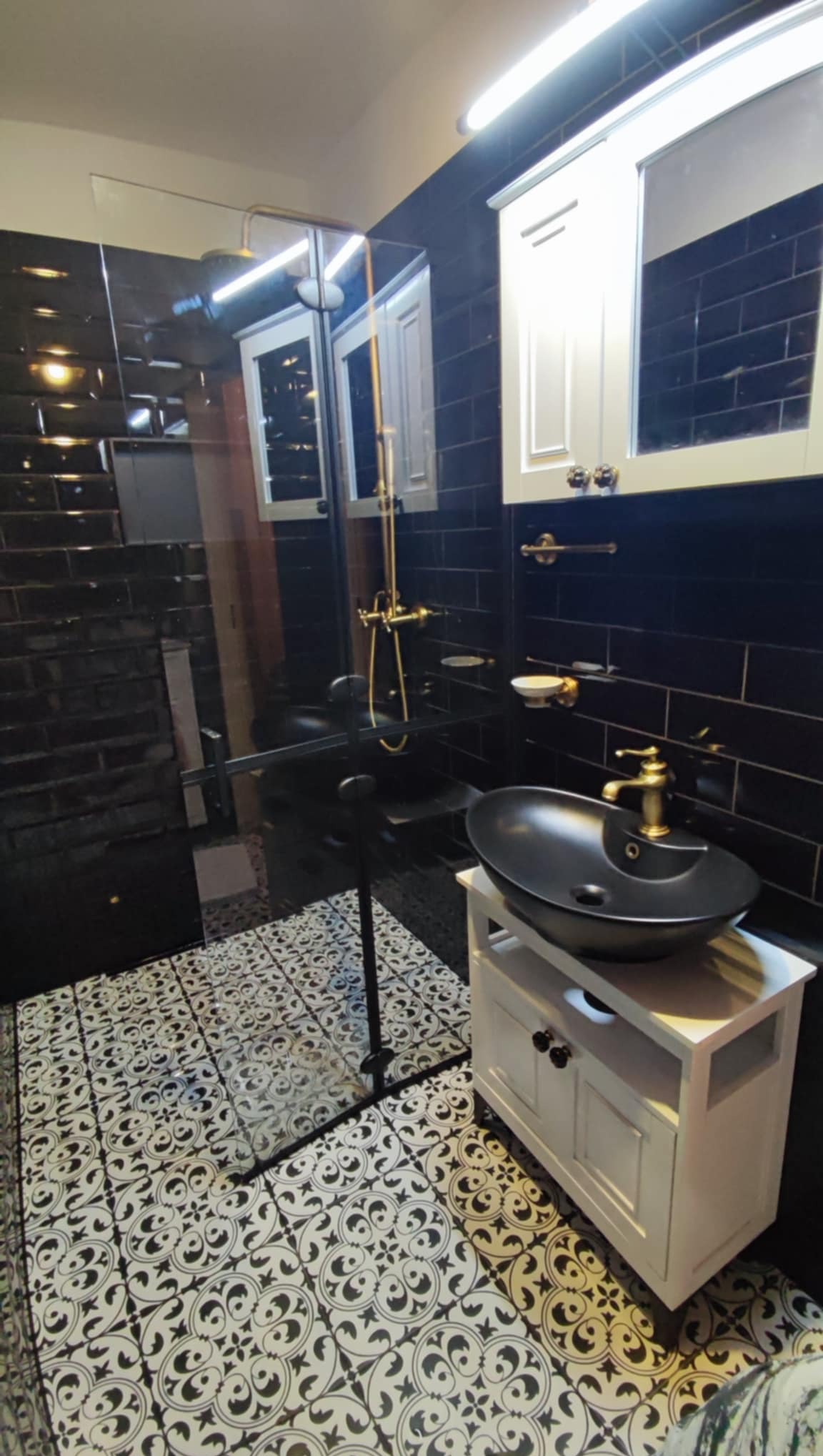 Elegancka łazienka w czerni i bieli z kabiną prysznicową, umywalką na szafce i złotą armaturą, wykończona błyszczącymi, czarnymi płytkami i wzorzystą podłogą.