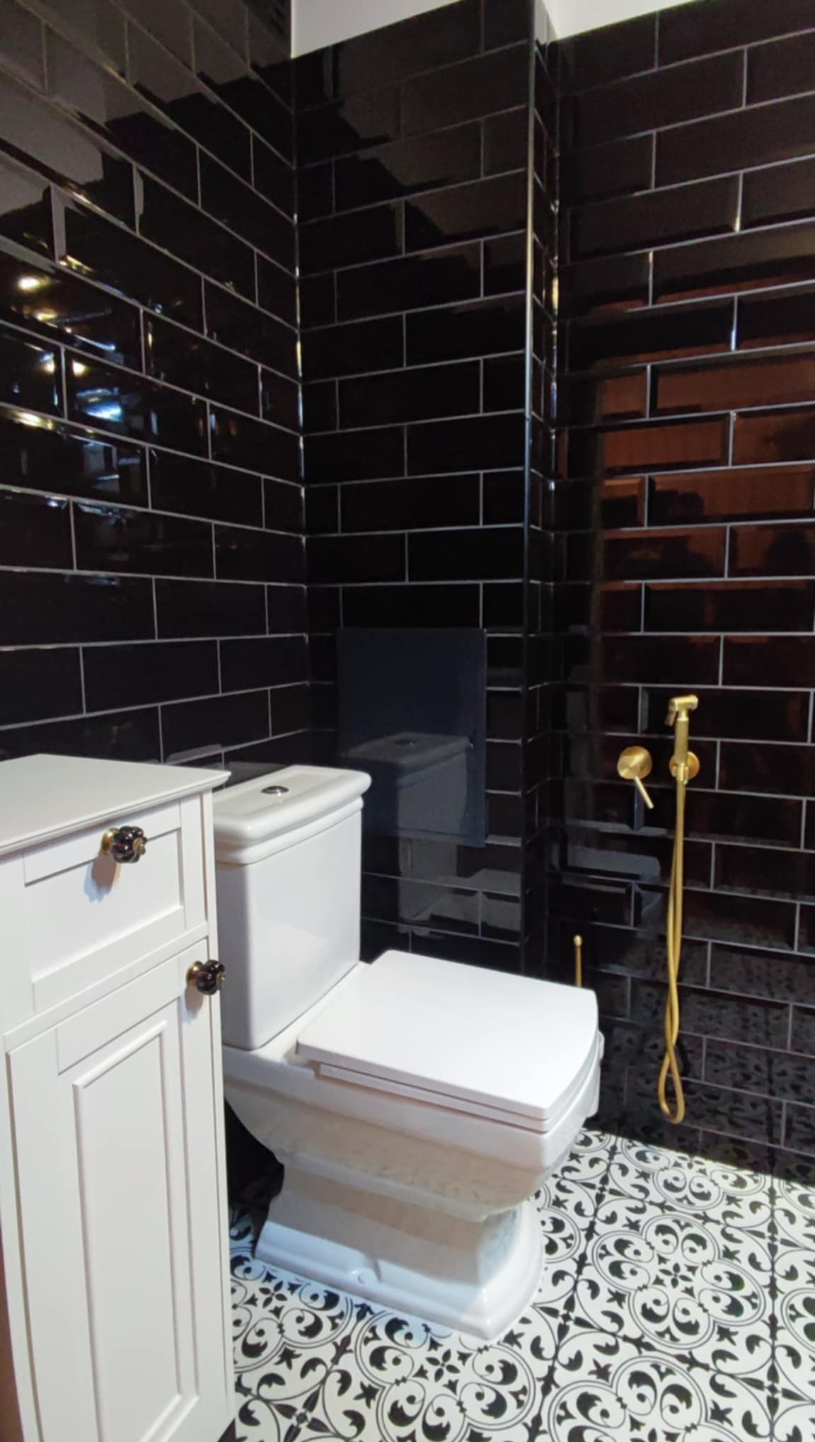 Nowoczesna łazienka z czarnymi, błyszczącymi płytkami, białą armaturą i szafką. Na podłodze wzorzyste, czarno-białe płytki. Złota słuchawka bidetowa.