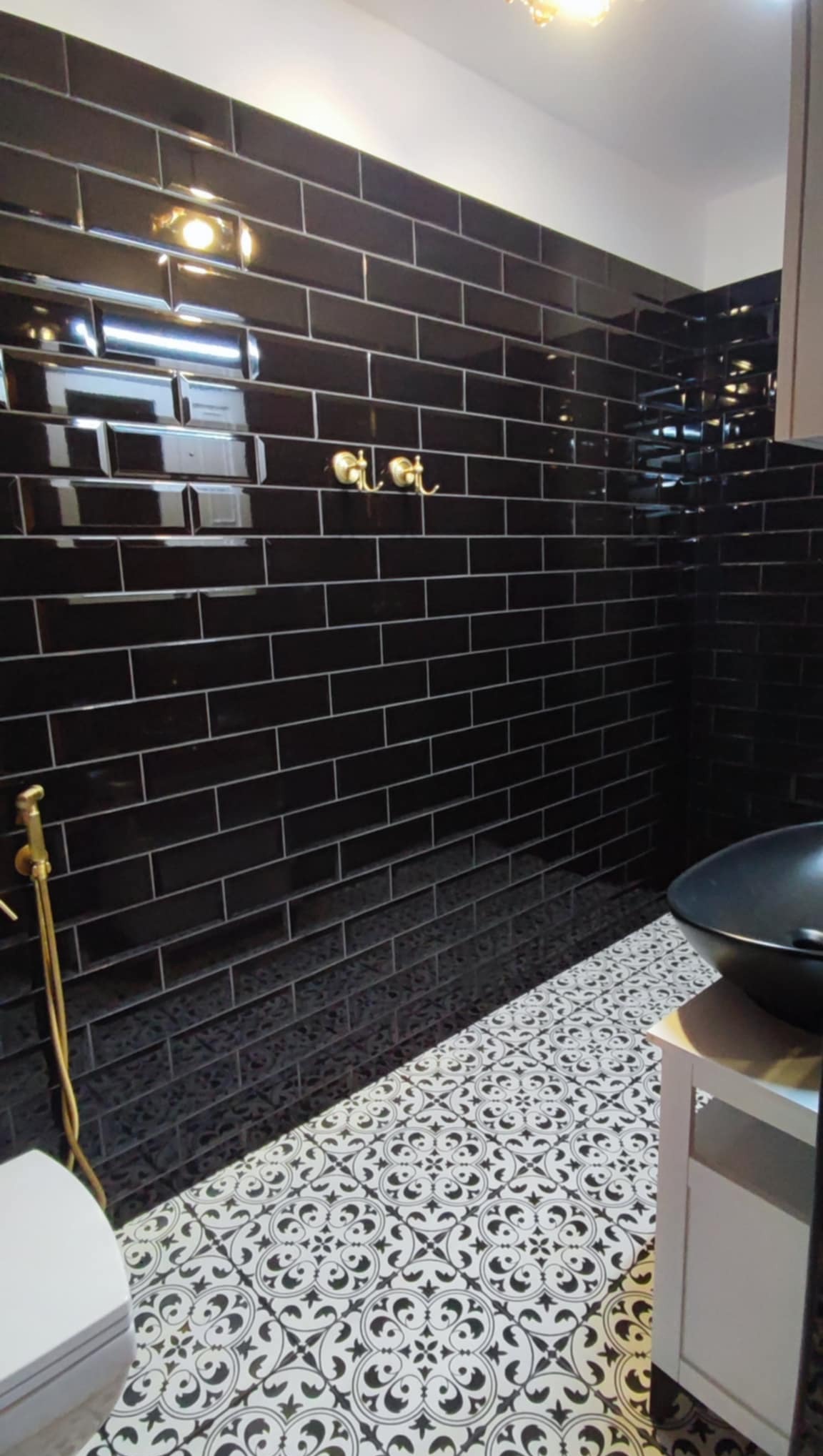 Elegancka łazienka z czarnymi, błyszczącymi płytkami na ścianach i wzorzystą, biało-czarną podłogą. Złote akcenty armatury i oświetlenia dopełniają luksusowy wystrój.