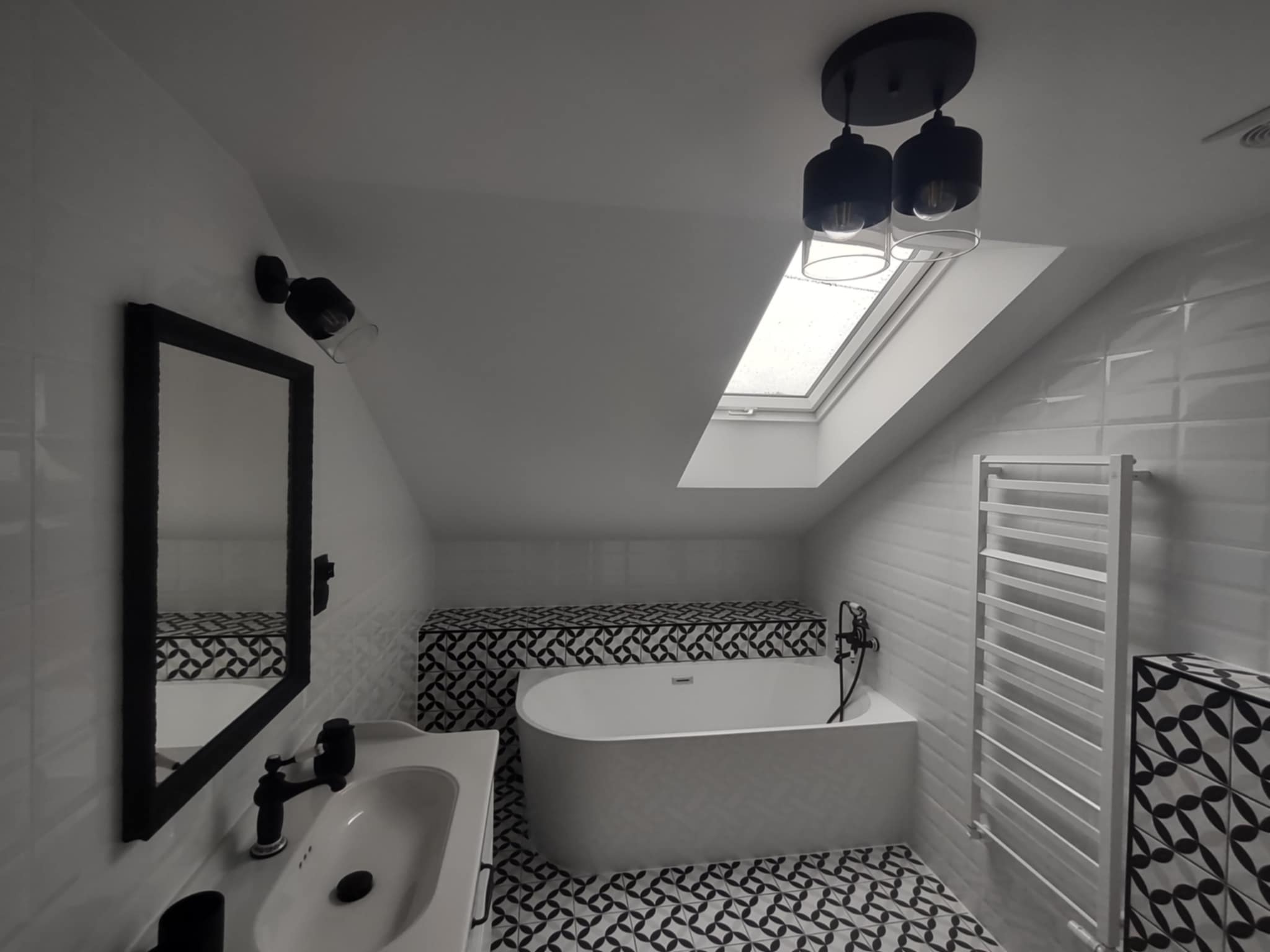 Nowoczesna łazienka na poddaszu z białą wanną, umywalką i czarną armaturą. Geometryczne wzory płytek na podłodze i obudowie wanny. Okno dachowe wpuszcza naturalne światło.