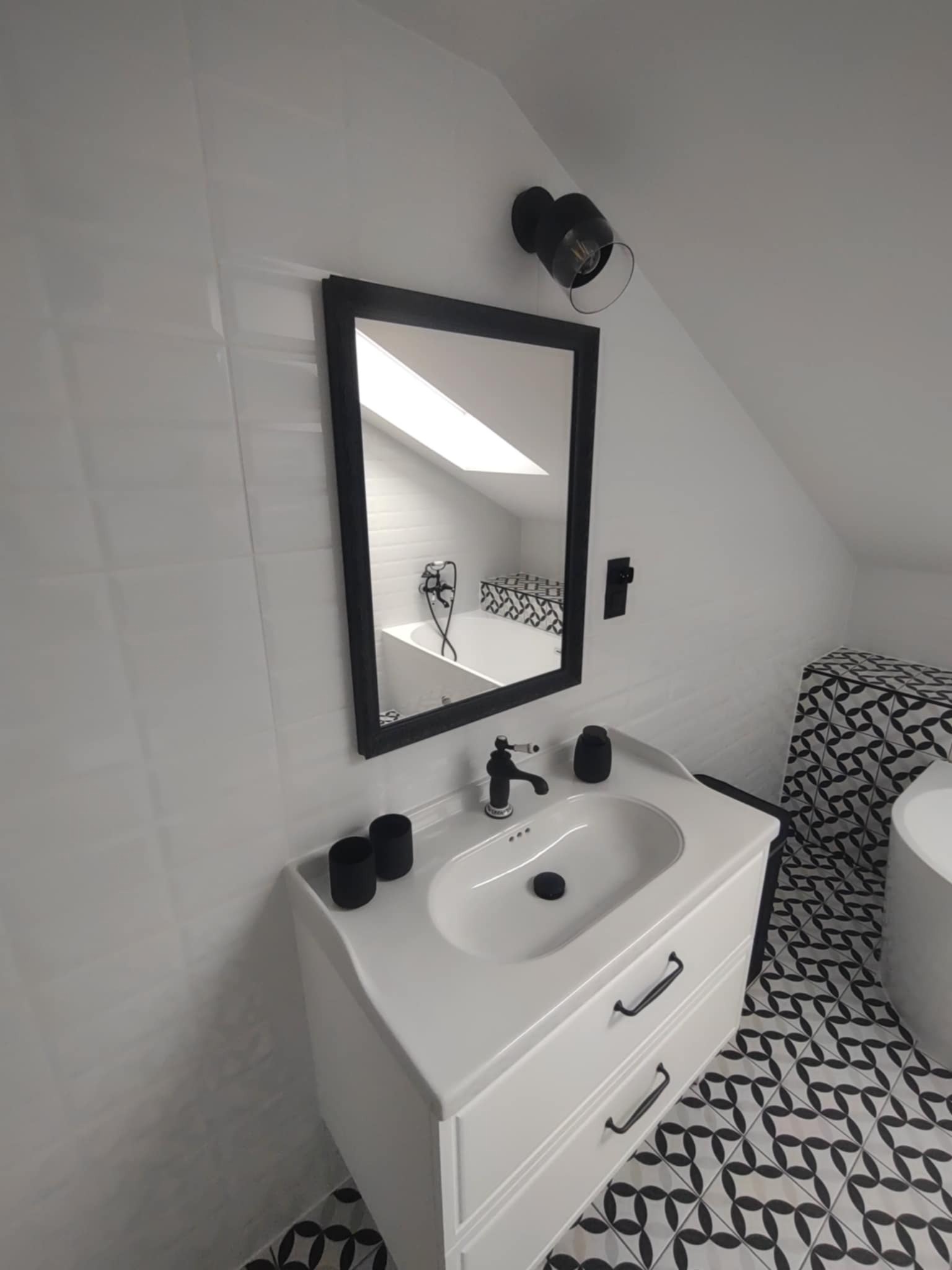Biała łazienka na poddaszu z umywalką na szafce, czarną armaturą i lustrem w czarnej ramie. W odbiciu widać wannę i okno dachowe. Uwagę przykuwają płytki podłogowe.