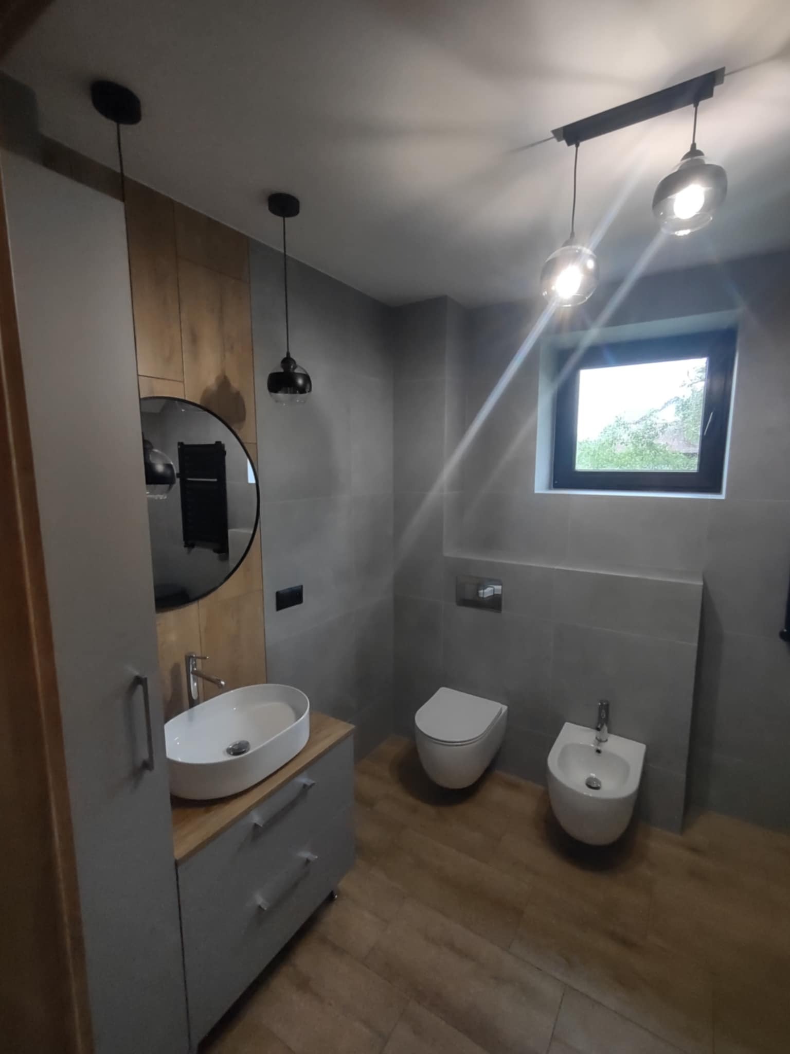 Nowoczesna łazienka w szarościach z drewnianymi akcentami. Umywalka nablatowa, okrągłe lustro, podwieszana toaleta i bidet. Oświetlenie punktowe i drewnopodobna podłoga.