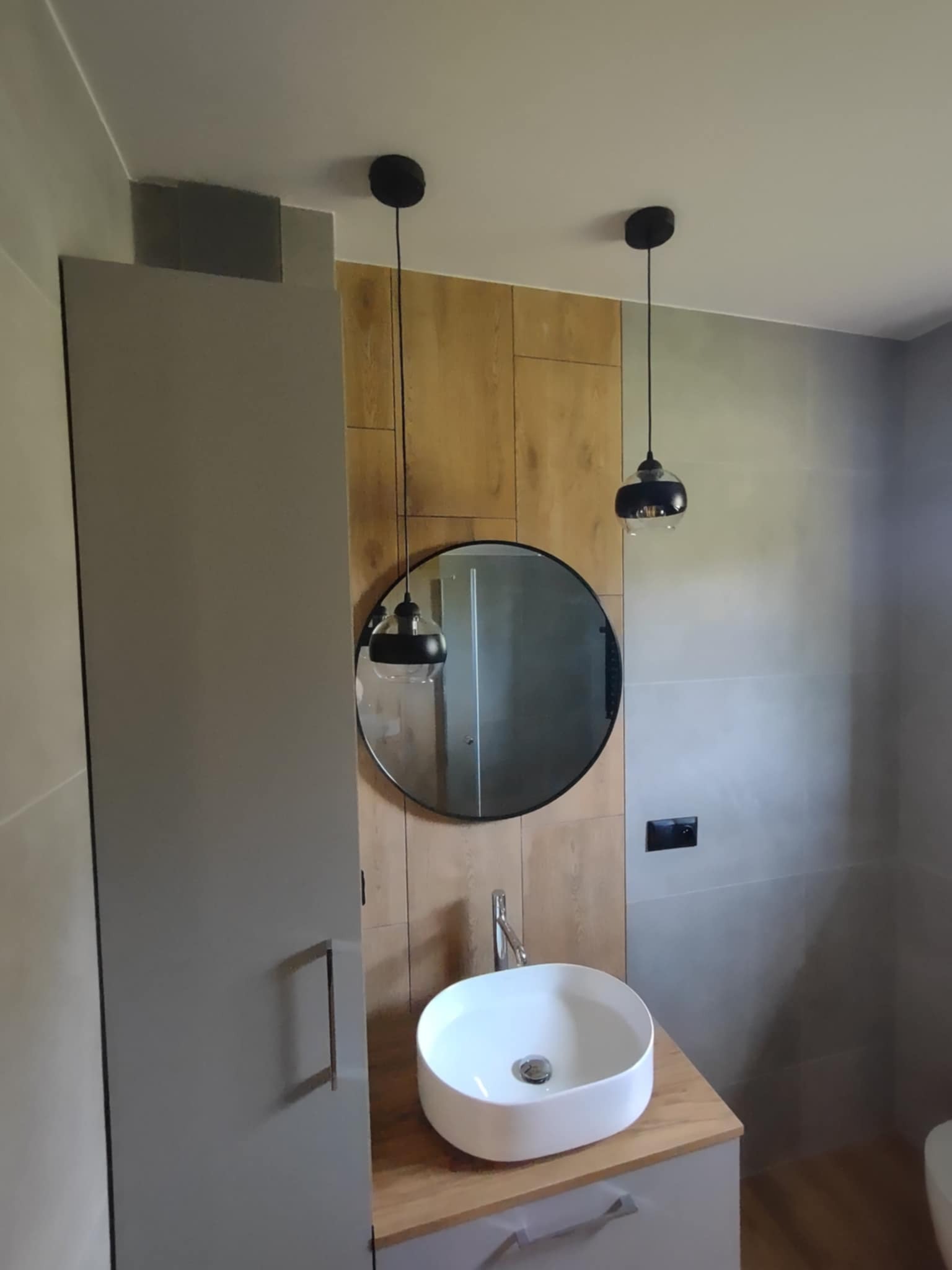 Stylowa łazienka po remoncie: szara szafka z umywalką, okrągłe lustro i drewnopodobne panele ścienne, oświetlone dwoma czarnymi lampami wiszącymi.