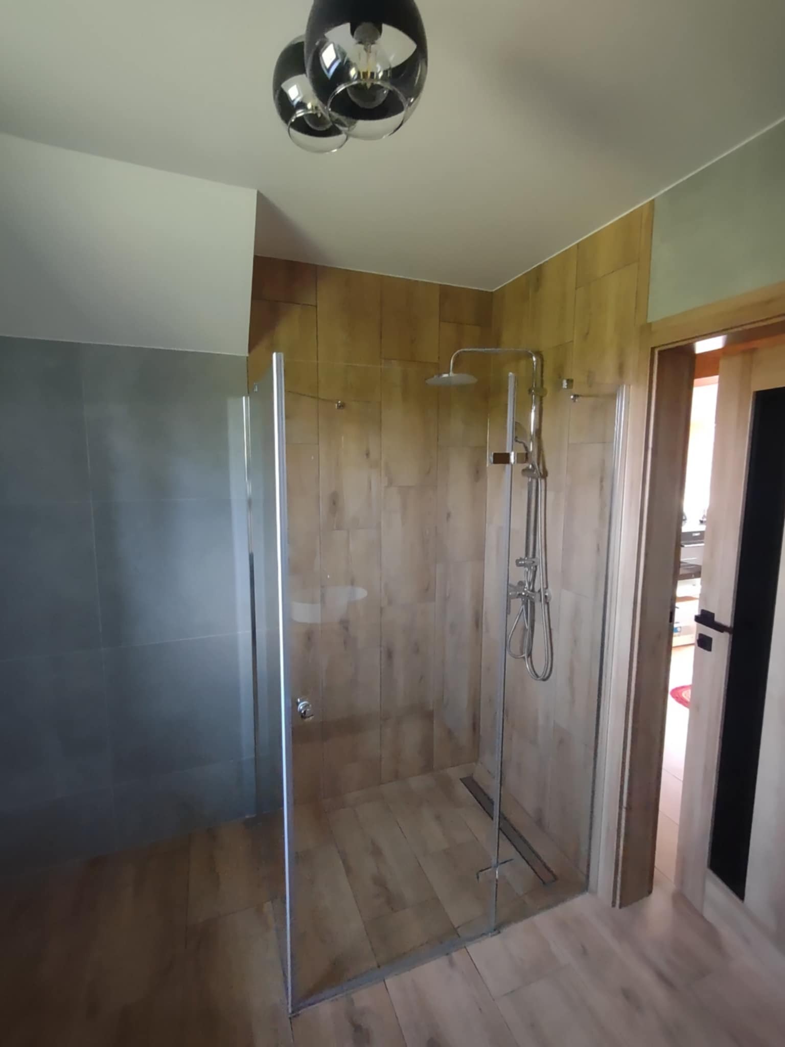 Kabina prysznicowa ze szklaną ścianką, wykończona płytkami imitującymi drewno, z widocznym odpływem liniowym i nowoczesną armaturą, w jasnej łazience.