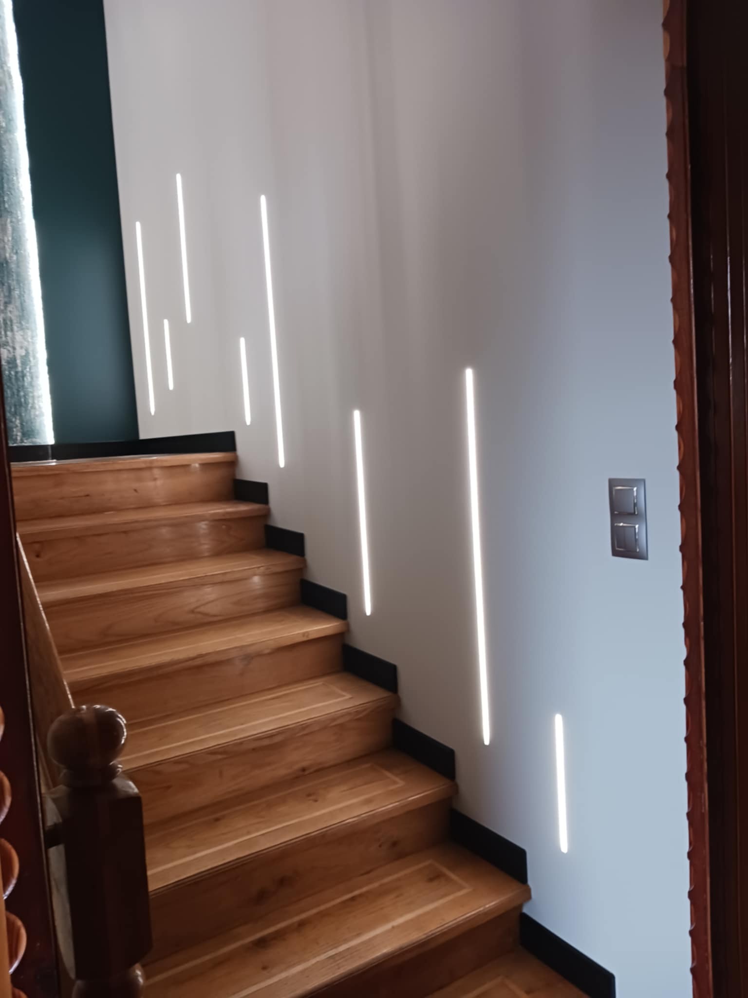Drewniane schody z podświetleniem LED na ścianie, widoczne detale wykończenia i estetyczne oświetlenie. Fragment drewnianych drzwi po prawej stronie kadru.
