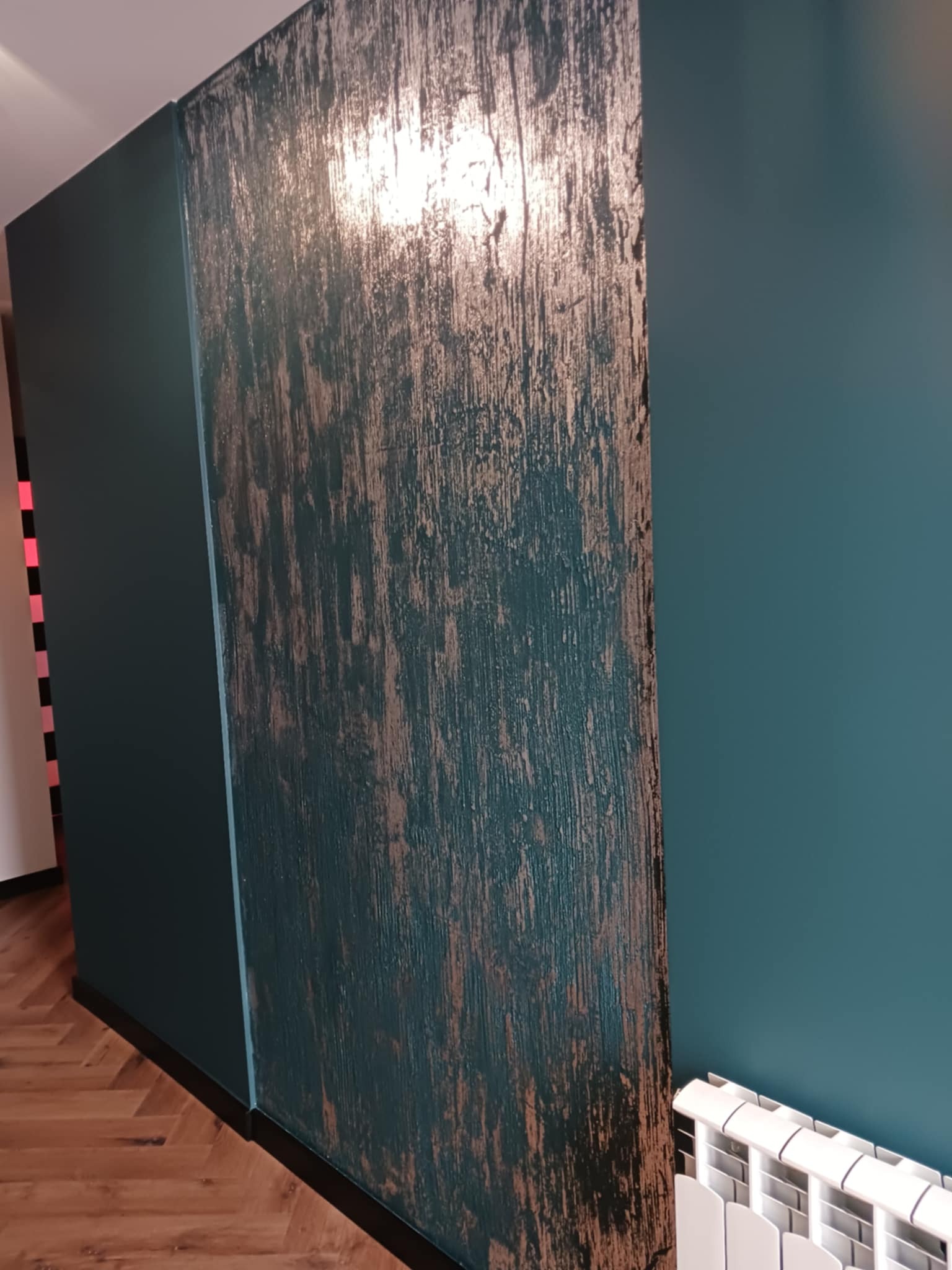 Stylowa ściana w odcieniach zieleni i brązu z unikatową fakturą,  w tle widoczny fragment parkietu i grzejnika, tworząc eleganckie wykończenie wnętrza.